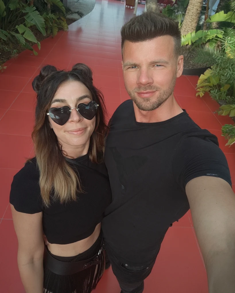 Paar steht lächelnd nebeneinander auf rotem Weg, umgeben von tropischen Pflanzen